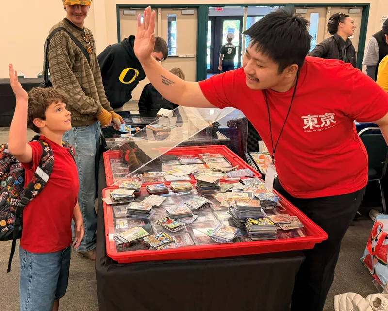 A kid high-fiving a vendor over a table of Pokémon cards
