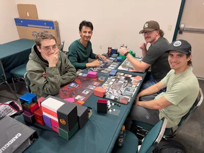 Four players mid-game at a Trade & Play table, decks, dice and deck boxes spread out